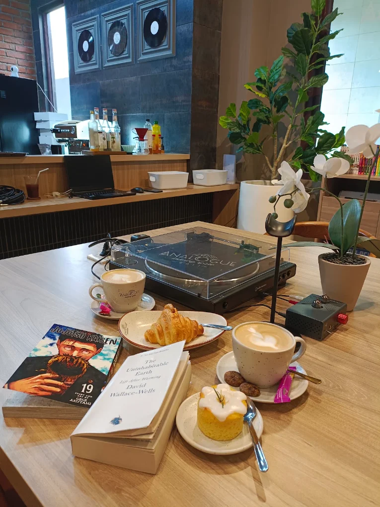 Meja baca dengan turntable, dua cangkir kopi, croissant, kue, dan dua buku.