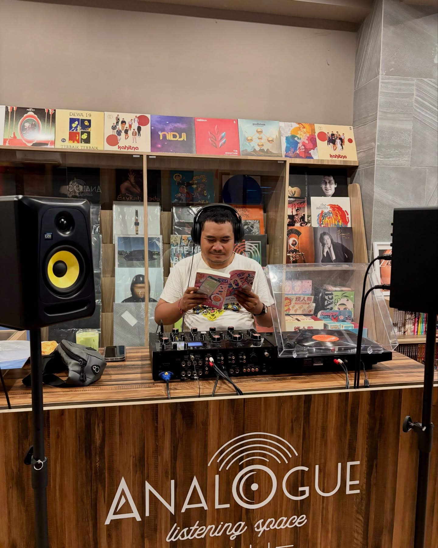 Peserta berheadphone membaca buku di balik meja DJ Analogue, dengan dinding sampul piringan hitam di belakangnya.