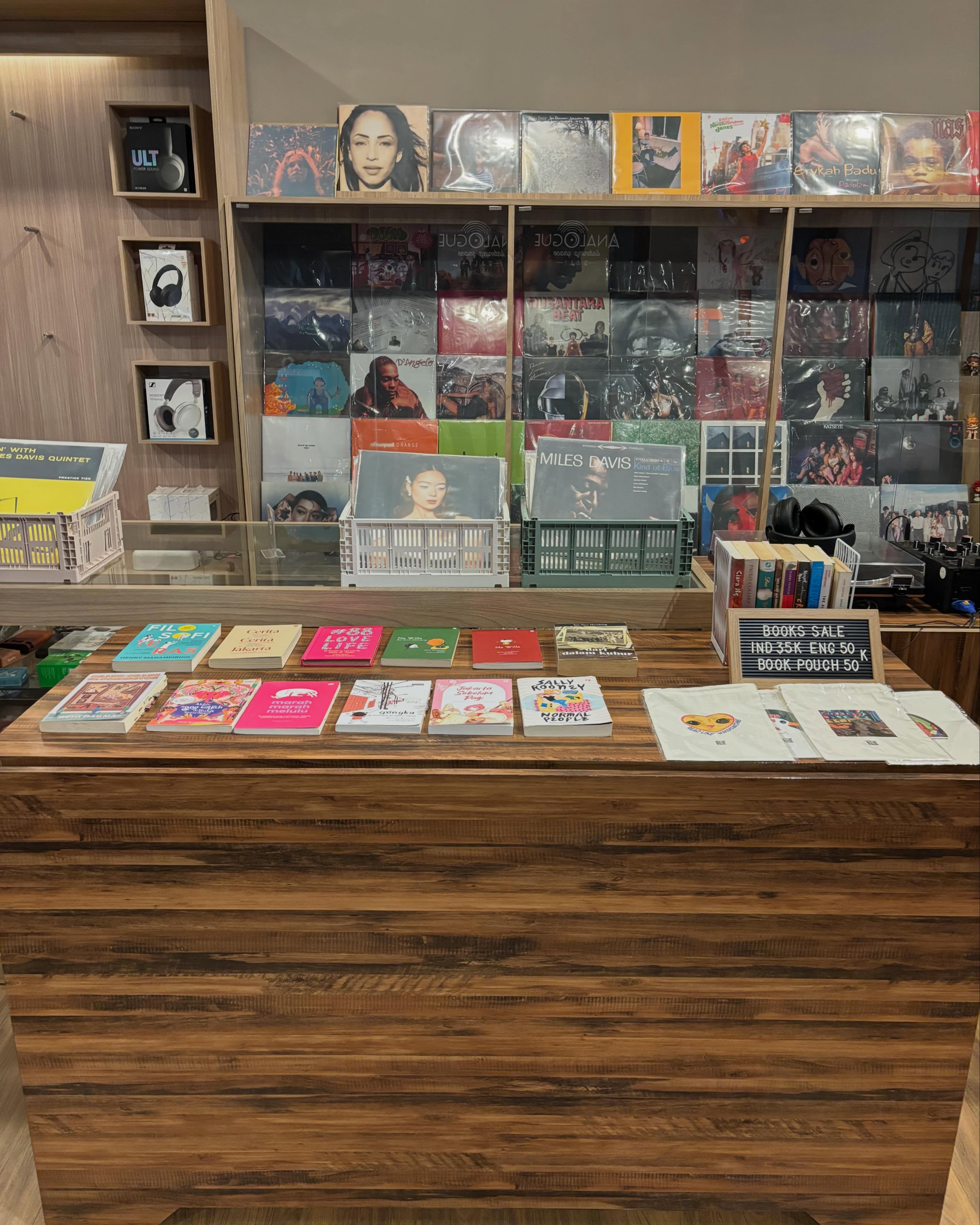 Meja kasir Analogue Listening Space: buku-buku dijajarkan di atas meja kayu, di depan dinding piringan hitam.