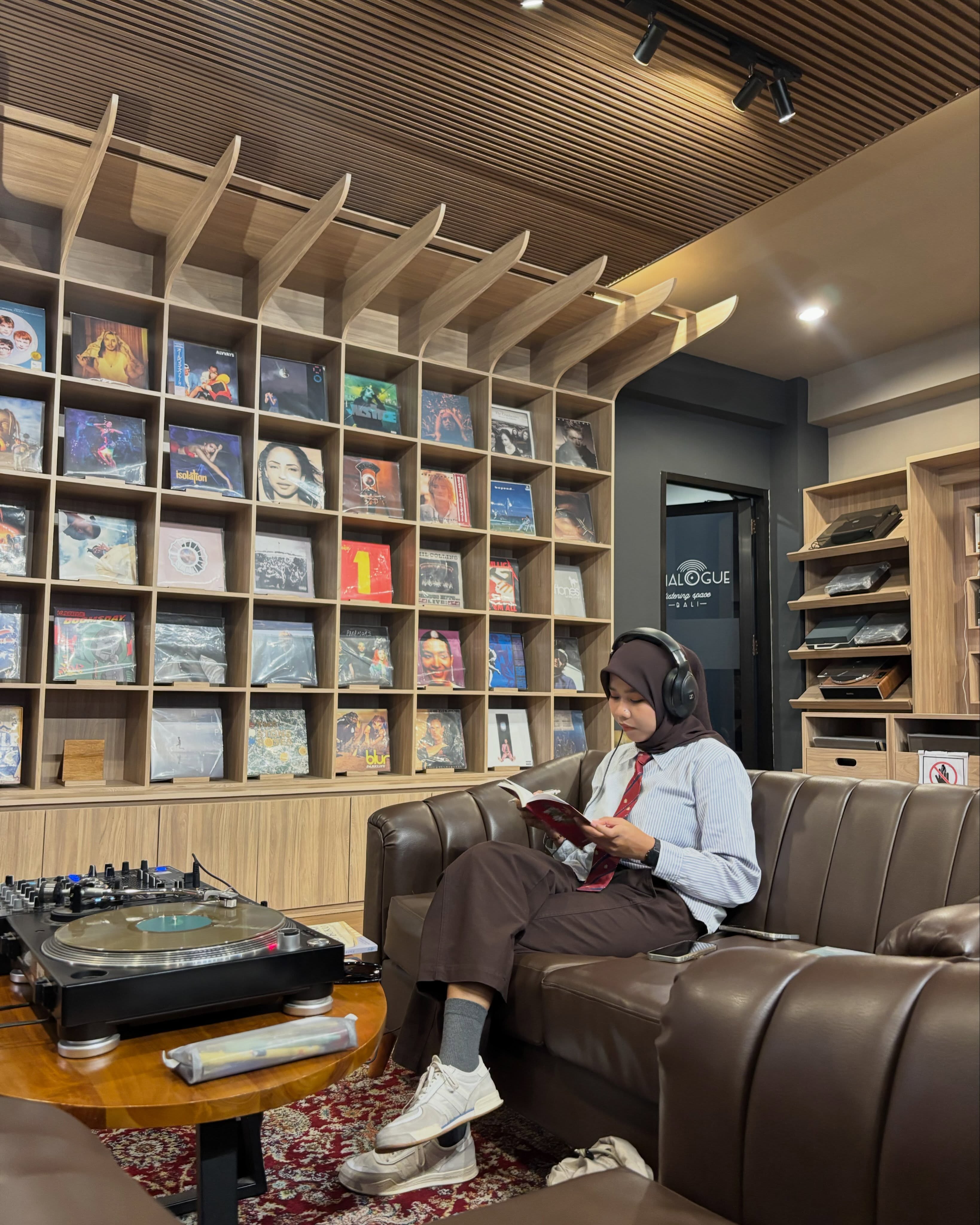 Peserta membaca sambil memakai headphone di sofa Analogue Listening Space, di depan rak berisi ratusan piringan hitam.